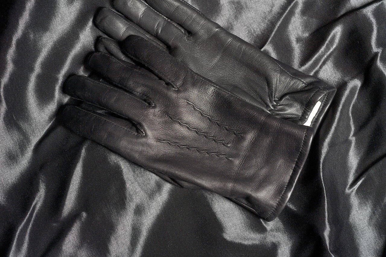 Pair of plain black leather gloves with stitched detailing, placed on a shiny black satin surface.

